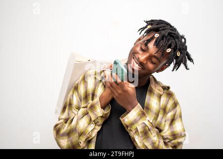 photo of young black guy on dreadlocks holding shopping bag, sending message on mobile phone. Happy African-American man using smartphone, enjoying ch Stock Photo