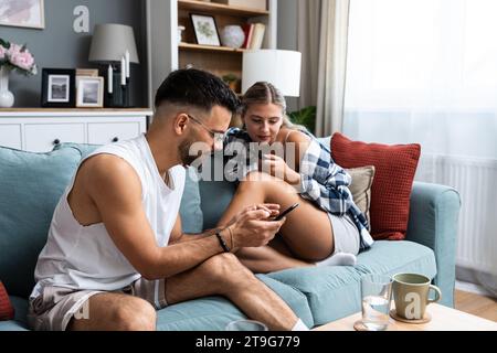 Young couple addicted to social network media scrolling their smartphones screens, spending free time together on sofa. Internet addiction concept. Stock Photo