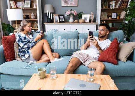 Young couple addicted to social network media scrolling their smartphones screens, spending free time together on sofa. Internet addiction concept. Stock Photo