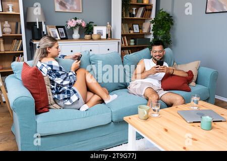 Young couple addicted to social network media scrolling their smartphones screens, spending free time together on sofa. Internet addiction concept. Stock Photo