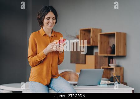Smiling woman texting on phone in a modern home office with a laptop Stock Photo