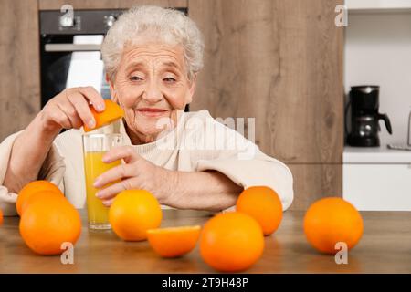 Senior woman squeezing orange juice into glass in kitchen Stock Photo