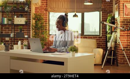 Freelancer enjoys working at workstation while listening to music and performing internet activities. At sunset, african american woman is multitasking, online career in blogging. Stock Photo