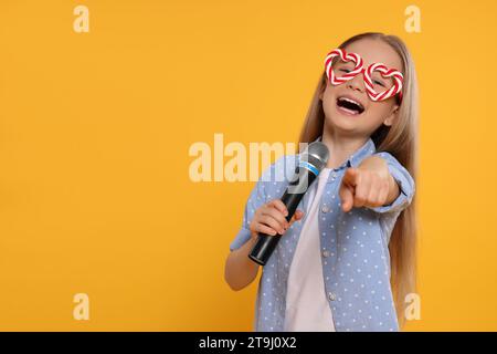 Cute little girl with funny glasses and microphone singing on yellow background, space for text Stock Photo