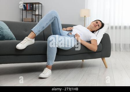 Young woman struggling to squeeze into tight jeans while lying on sofa at home Stock Photo