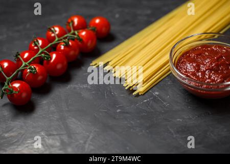 Cherry tomatoes branch, raw spaghetti, tomato sauce - ingredients for cooking Italian pasta, on dark background, selective focus. Stock Photo
