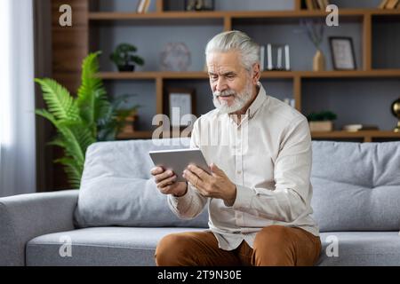 Senior gray-haired man sitting on sofa at home and using tablet, reading news, watching video. Stock Photo