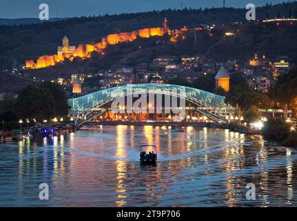 Night view along Mtkvari Kura river in capital of Georgia Tbilisi Tiflis city with a motor boat moving towards brightly lit Bridge of Peace in Stock Photo