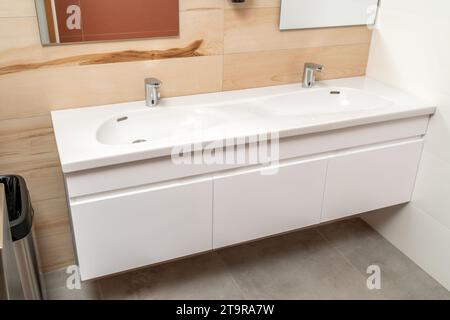 Wall mounted bathroom vanity cabinet with double white porcelain sink and silver sensor faucets in modern bathroom with concrete and wood tiles Stock Photo