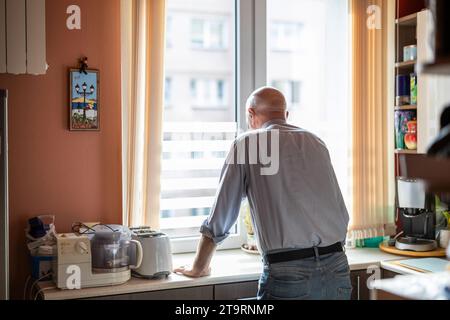 Senior man standing at the kitchen counter in his house and looking out the window Stock Photo