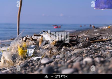 Bunch of collected cellophane bags, plastic on the seashore on a pebble beach in the background people are swimming in disfocus. Concept of ecology Stock Photo