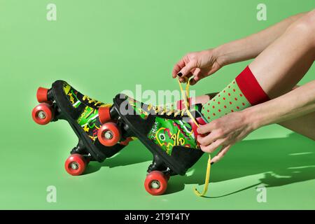 Side view of crop anonymous female tying laces and put on colorful retro roller blades against green wall while trying roller skating Stock Photo