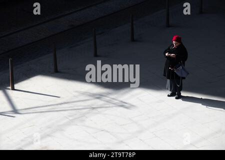 Belgrade, Serbia- March 18, 2022: One elderly woman with red hat standing alone on a town square street on the cold sunny day, high angle view Stock Photo
