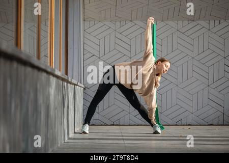 Attractive young woman practicing yoga, performing Trikonasana exercise, triangle pose, outdoors, working out with a fitness belt in sportswear, stand Stock Photo