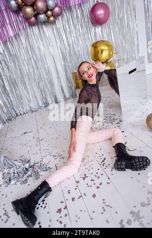 Fashion girl with beauty makeup, in soft pink pants, mesh bodysuit sits on floor among confetti, silver shiny foil tinsel. Close-up portrait of model Stock Photo