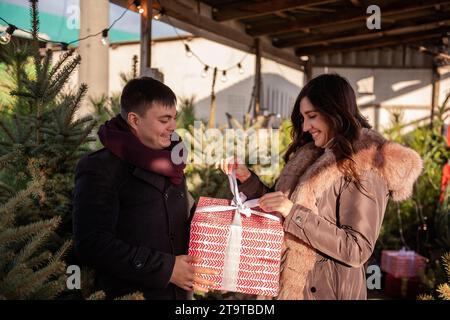 Close-up portrait of couple in love. Young man gives large red box to beautiful woman among the green Christmas tree market. Girl is surprised by unex Stock Photo