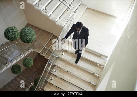 High angle shot of African American businessman wearing suit carrying laptop descending stairs holding on banister in modern office block, copy space Stock Photo