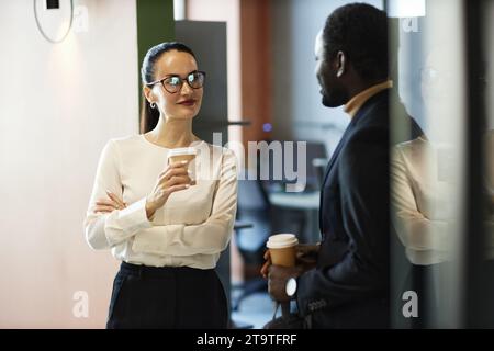 Medium shot of diverse colleagues chatting and standing in office building hall during coffee break, focus on businesswoman, copy space Stock Photo