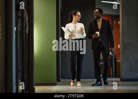 Full length shot of two cheerful diverse colleagues carrying gadgets talking and walking through office block, copy space Stock Photo