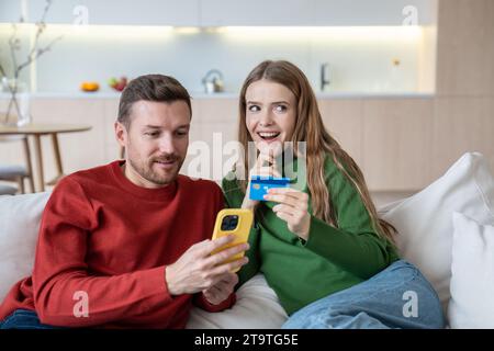 Happy man woman doing online payment in smartphone sitting on couch at home. Stock Photo