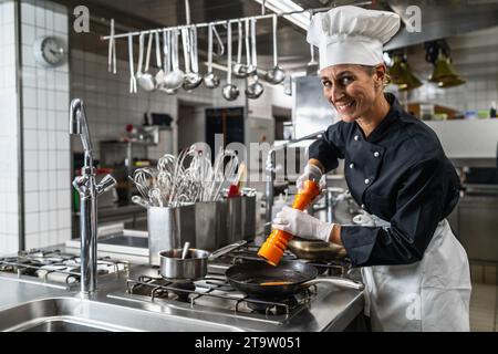 Smiling female chef in hotel or restaurant kitchen cooking and seasoning the food with a pepper or salt mill Stock Photo