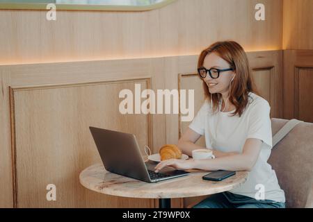Focused female student attending an online lecture in a cafe, with laptop, coffee, and pastry. Stock Photo