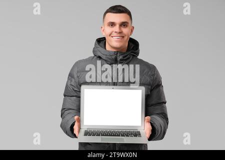 Young man in stylish puffer jacket with modern laptop on grey background Stock Photo