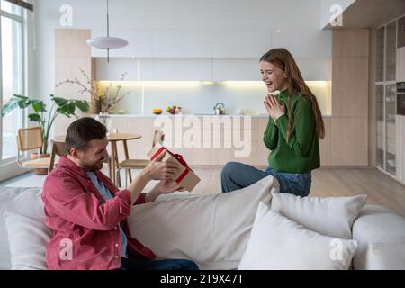 Laughing woman sitting on sofa, looking with excitement as beloved man trying to unwrap giftbox Stock Photo