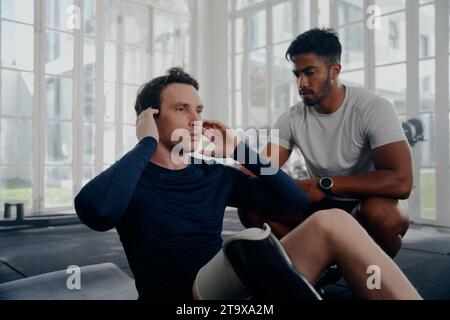 Multiracial trainer next to young man in sportswear with artificial limb doing push-ups at the gym Stock Photo