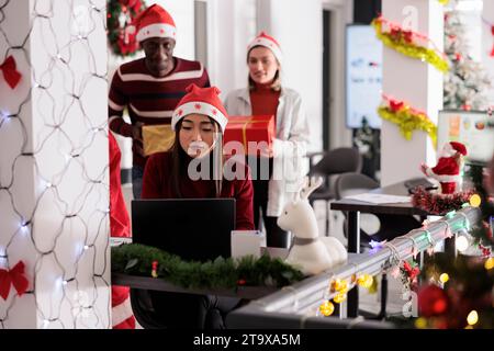 Coworkers in xmas ornate office teaming up to surprise unsuspecting employee focused on finishing project. Corporate team sneaking behind colleague with Christmas presents during holiday season Stock Photo