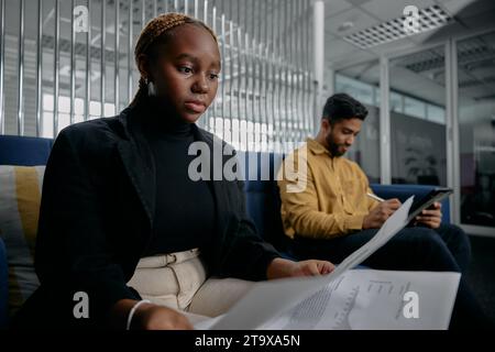 Two multiracial young adults in businesswear reading documents on sofa in corporate office Stock Photo
