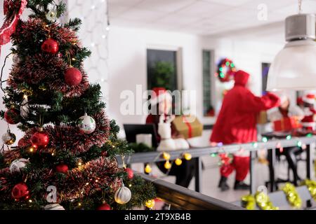 Festively decorated christmas tree close up with business workers receiving presents from Santa Claus in blurry background. Xmas adorn modern workspace during winter holiday season Stock Photo