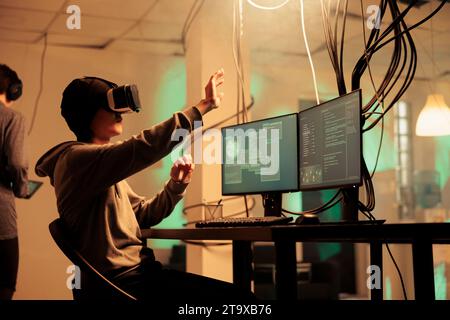 Male hacker using vr glasses to hack computer system, planting trojan virus for malware at night. Young adult working with 3d virtual reality on headset, modern technology hacking. Stock Photo