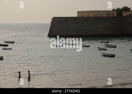 Beach goers wade into the water by the small fishing boat harbor along the walls of the Castillo de Santa Catalina in Cadiz, Spain. The public beach sits between two ancient castles on the bay. Stock Photo