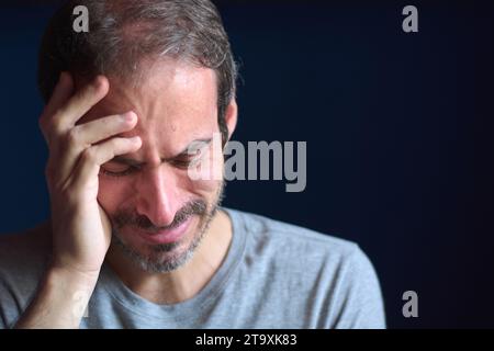 Close up of young caucasian 30 year old man with short beard and blue eyes crying while holding his head. Concept of depression, loss and grief. Emoti Stock Photo