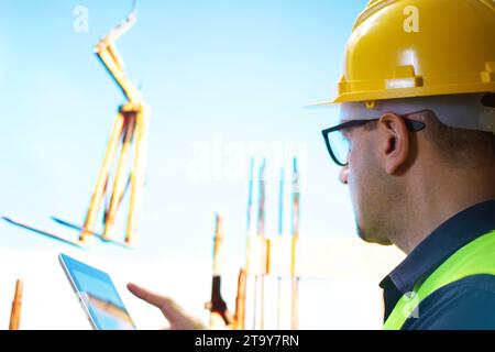 Back view of caucasian engineer standing on construction site checking plan on digital tablet. Stock Photo