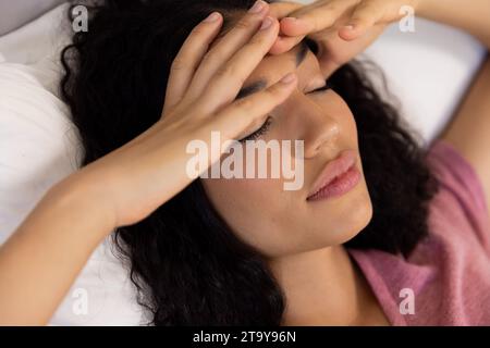 Tired biracial woman holding hands on her head lying in bed at sunny home Stock Photo
