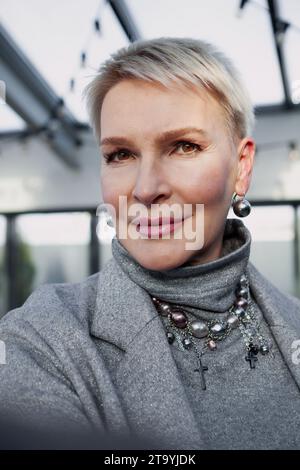 Selfie of beautiful mature blonde woman with short haircut. Close up portrait of stylish middle aged female model with brown eyes looking into the cam Stock Photo