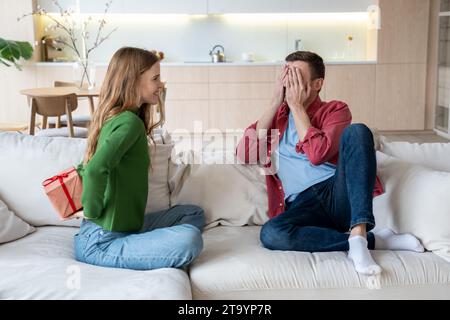Cheerful girlfriend sitting on sofa hiding box behind and intrigued boyfriend waiting for present Stock Photo