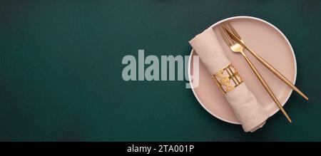 Plate with fork and knife. Mockup with empty space. Gold cutlery, dark green background. Top view. Celebration place setting, fabric napkin. Dining ta Stock Photo