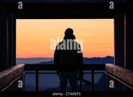 A man standing in an elevated position, gazing out over a mountainous landscape at sunset Stock Photo