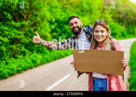 Happy couple hitchhiking on roadside trying to stop car. They are holding blank cardboard for your text. Stock Photo