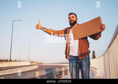 Man is hitchhiking on roadside trying to stop car. He is holding blank cardboard for your text. Stock Photo