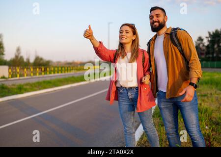 Happy couple hitchhiking on roadside trying to stop car. Stock Photo