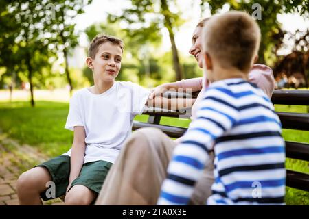 Happy mother is sitting with her sons on bench in park. They are talking and enjoying their time together. Stock Photo