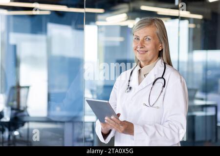 Portrait of a smiling senior female doctor standing in a clinic in a white coat with a stethoscope, holding a tablet and looking at the camera. Stock Photo