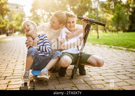 Joyful mother having fun with her two sons in park on sunny day. They are ridding skateboard and scooter. Stock Photo