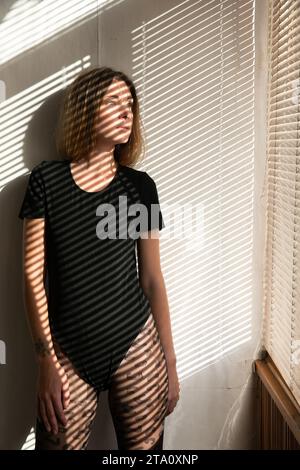 A woman stands at the window in the shade of the blinds Stock Photo