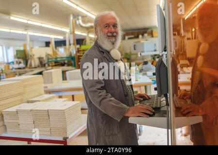 Smiling bearded man operating a a cutting machine computer in a carpentry workshop Stock Photo