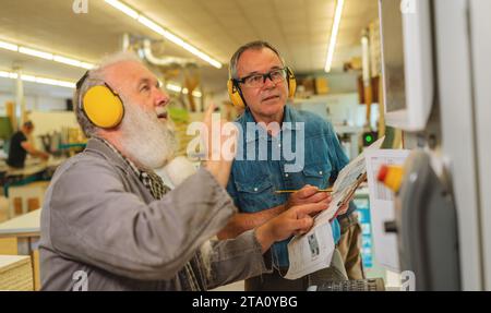 Two men with ear protection discussing work on a cutting machine in a busy carpentry workshop Stock Photo